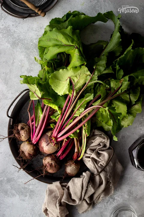 A bunch of whole beetroot complete with leaves covered in soil on a dark grey metal tray with a light brown linen napkin resting at the side. There is also an open jar of pickled beetroot, an empty pickling jar and a couple of stacked grey metal plates with a wooden fork on top in the background. Set over a brushed grey backdrop.