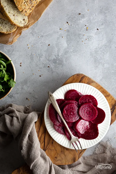 Close up view of a white plate filled with pickled beetroot topped with a fork, on top of a wooden chopping board. Set over a brushed grey backdrop there is also a light brown linen napkin, a bowl of salad leaves and some sliced bread in the background.