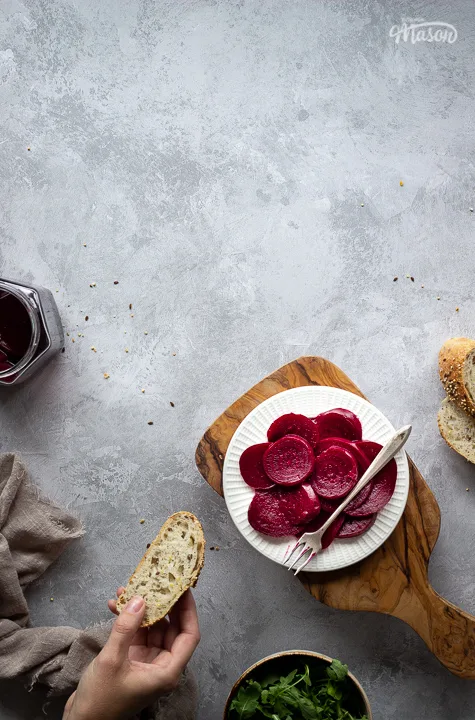 A white plate filled with pickled beetroot topped with a fork, on top of a wooden chopping board. Set over a brushed grey backdrop there is also an open jar of pickled beetroot, a light brown linen napkin, a bowl of salad leaves, some sliced bread and a hand holding a piece of bread in the background.