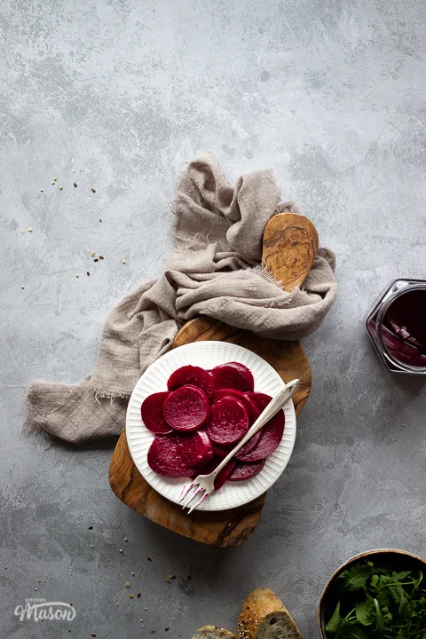 A white plate filled with pickled beetroot topped with a fork, on top of a wooden chopping board. Set over a brushed grey backdrop there is also an open jar of pickled beetroot, a light brown linen napkin, a bowl of salad leaves and some sliced bread in the background.