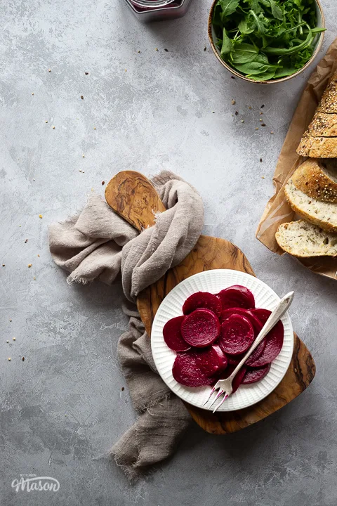A white plate filled with pickled beetroot topped with a fork, on top of a wooden chopping board. Set over a brushed grey backdrop there is also an open jar of pickled beetroot, a light brown linen napkin, a bowl of salad leaves and some sliced bread in the background.