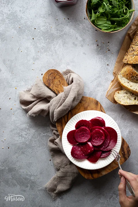 A white plate filled with pickled beetroot with a hand reaching in with a fork to take a piece, on top of a wooden chopping board. Set over a brushed grey backdrop there is also an open jar of pickled beetroot, a light brown linen napkin, a bowl of salad leaves and some sliced bread in the background.