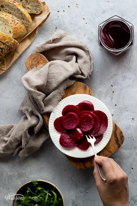 A white plate filled with pickled beetroot with a hand reaching in with a fork to take a piece, on top of a wooden chopping board. Set over a brushed grey backdrop there is also an open jar of pickled beetroot, a light brown linen napkin, a bowl of salad leaves and some sliced bread in the background.