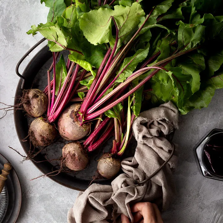 Close up of a bunch of whole beetroot complete with leaves covered in soil on a dark grey metal tray with a light brown linen napkin resting at the side and a hand holding it. There is also an open jar of pickled beetroot and a couple of stacked grey metal plates with a wooden fork on top in the background. Set over a brushed grey backdrop.