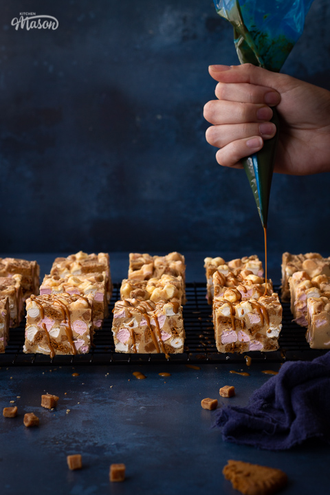 Distanced front view of melted Biscoff spread being drizzled over Biscoff rocky road bars set on a wire cooling rack. Set on a blue backdrop, there is also a blue linen napkin and some fudge pieces scattered in the background.