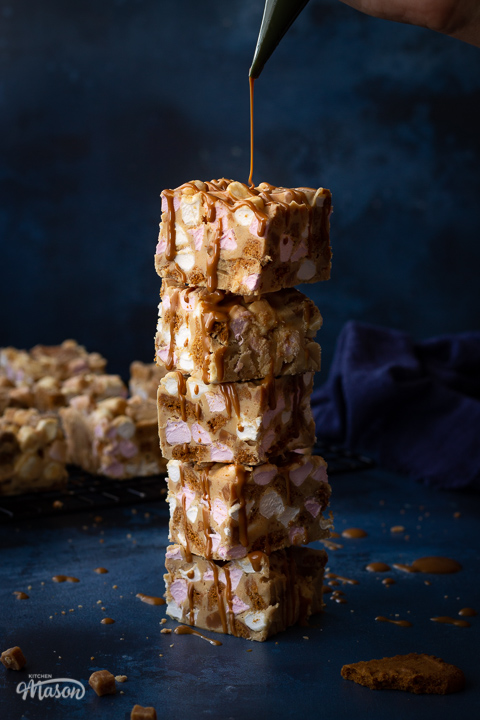 Front view of a stack of 5 Biscoff rocky road bars being drizzled with melted Biscoff spread. Set on a blue backdrop, there is also more Biscoff rocky road bars on a wire rack, a blue linen napkin, broken Biscoff biscuits and fudge pieces scattered in the background.