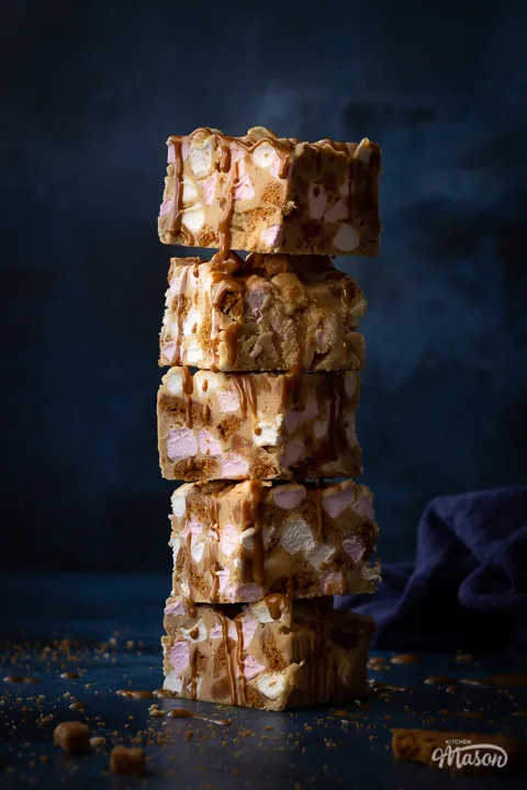A close up front view of a stack of 5 Biscoff rocky road bars that have been drizzled in more Biscoff spread. Set on a blue backdrop, there's also a blue linen napkin, broken Biscoff biscuits and fudge pieces scattered in the background.