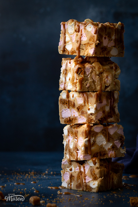 A close up front view of a stack of 5 Biscoff rocky road bars that have been drizzled in more Biscoff spread and stacked up to the side. Set on a blue backdrop, there's also a blue linen napkin, broken Biscoff biscuits and fudge pieces scattered in the background.