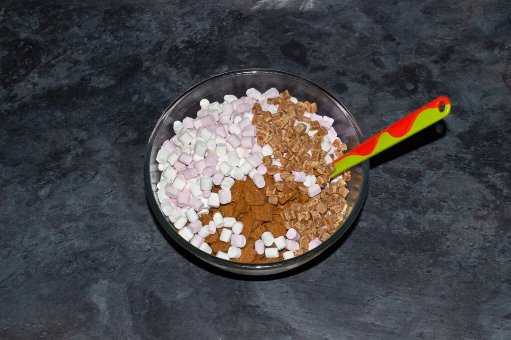 A large glass bowl on a kitchen worktop filled with a melted white chocolate and Biscoff mixture, mini marshmallows, chopped Biscoff biscuits and fudge pieces.