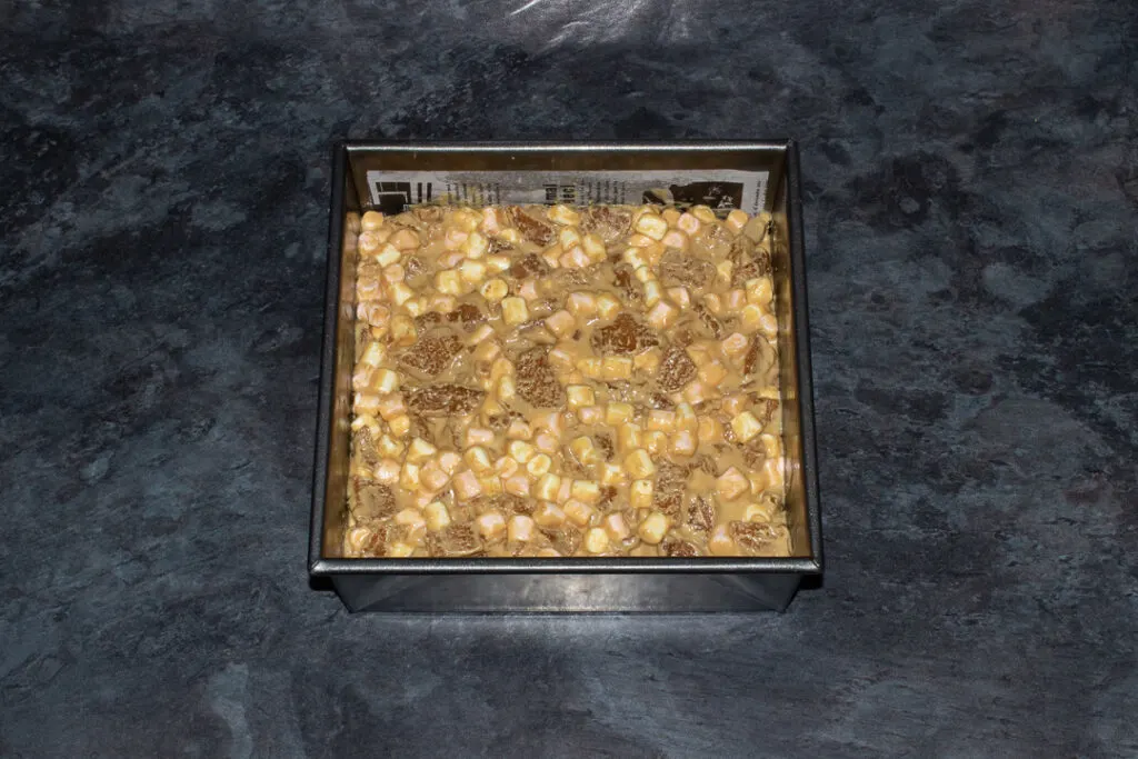 A lined square baking tin on a kitchen worktop filled with Biscoff rocky road.