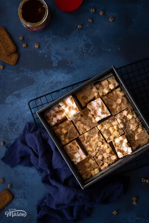 A square baking tin filled with bars of Biscoff rocky road set on a wire cooling rack with a blue linen napkin. There is also an open jar of Biscoff spread, some Biscoff biscuits and some fudge pieces scattered around in the background. All set on a deep blue backdrop.