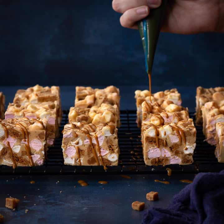 Close up front view of melted Biscoff spread being drizzled over Biscoff rocky road bars set on a wire cooling rack. Set on a blue backdrop, there is also a blue linen napkin and some fudge pieces scattered in the background.