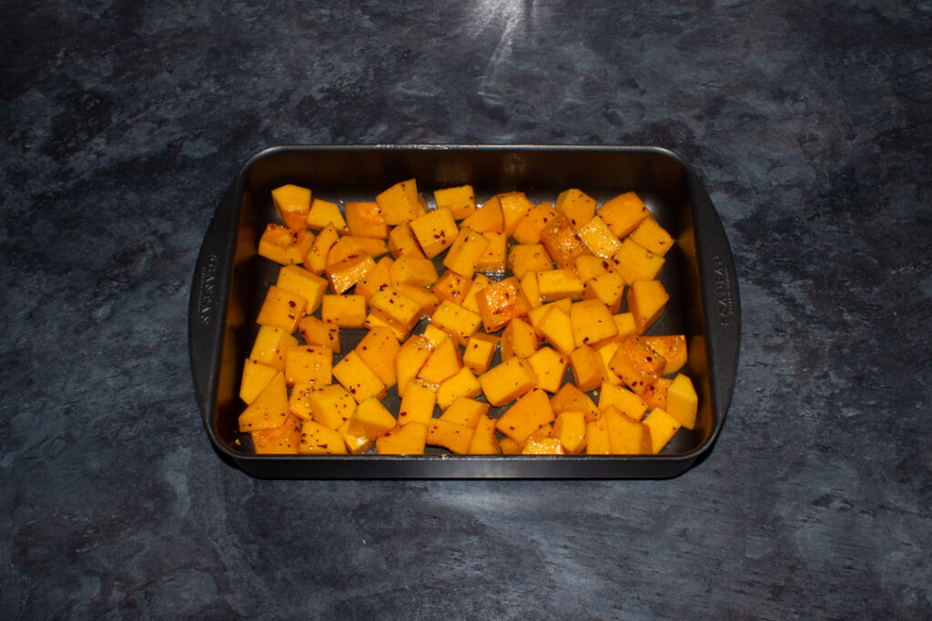 Butternut squash cubes in a roasting pan mixed with oil, chilli flakes and seasoning. Set on a kitchen worktop.