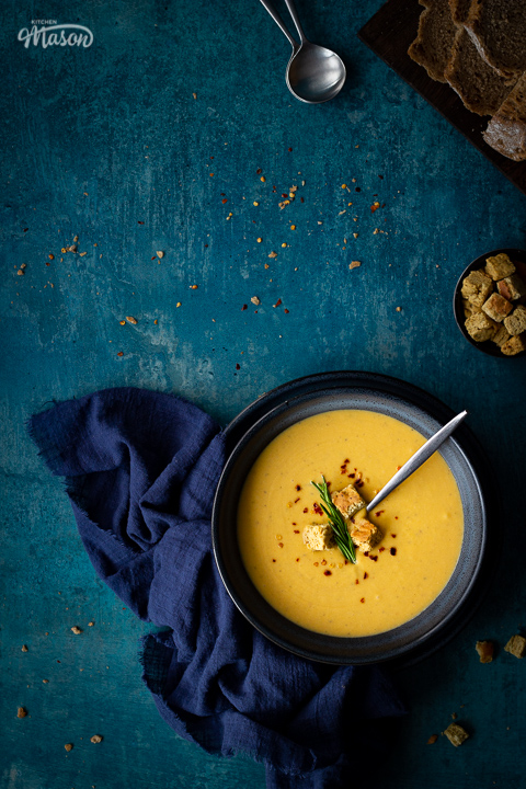 Flat lay view of spicy roast butternut squash soup with a spoon in a blue bowl set on a blue plate with a dark blue linen napkin on the side. Set on a teal backdrop, there's also a board with sliced bread, a pot with croutons and two soup spoons in the background.