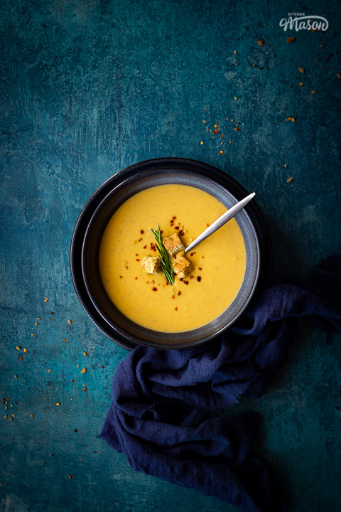 Flat lay view of spicy roast butternut squash soup with a spoon in a blue bowl set on a blue plate with a dark blue linen napkin on the side. Set on a teal backdrop, there are a few chilli flakes and breadcrumbs scattered in the background.