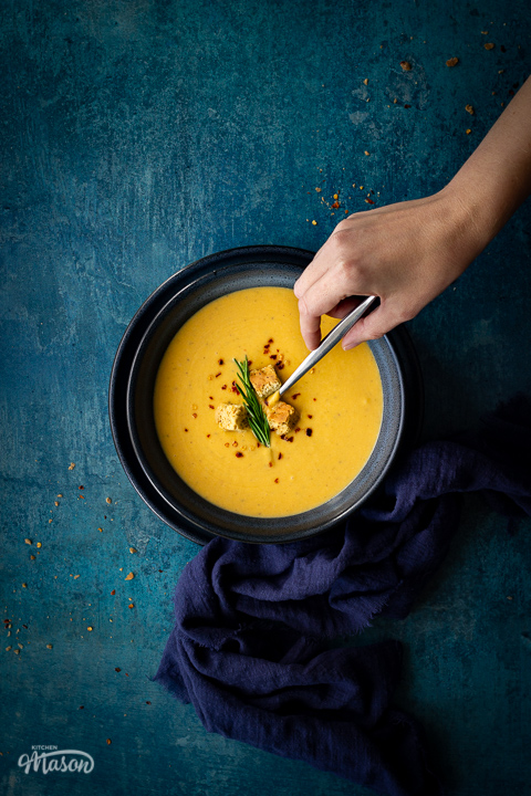 Flat lay view of spicy roast butternut squash soup with a hand reaching for a spoon in a blue bowl set on a blue plate with a dark blue linen napkin on the side. Set on a teal backdrop, there are a few chilli flakes and breadcrumbs scattered in the background.