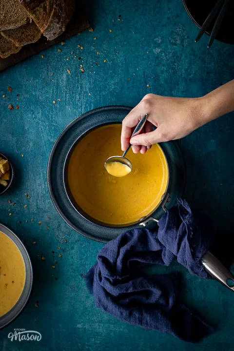 Flat lay view of a saucepan filled with spicy roast butternut squash soup and a hand reaching in with a spoon set on a blue plate with a deep blue linen napkin on the side. Set on a teal backdrop, there is also a bowl of soup, a pot of croutons, a board with sliced bread and stacked bowls with soup spoons inside in the background.