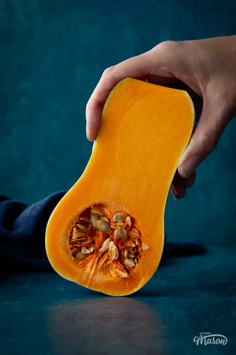 Close up front view of someone holding half a butternut squash against a teal backdrop. There is a dark blue linen napkin scrunched up in the background.