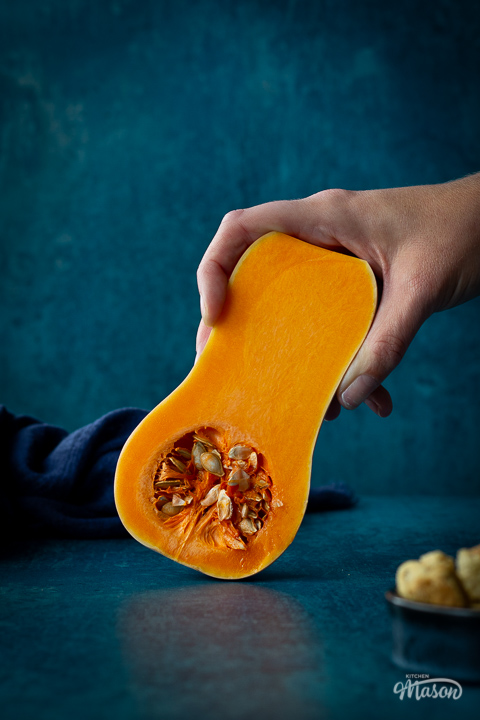 Distanced front view of someone holding half a butternut squash against a teal backdrop. There is a dark blue linen napkin scrunched up and a pot of croutons in the background.