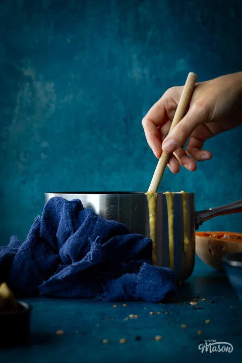 Someone stirring with a wooden spoon in a saucepan of butternut squash soup set against a teal backdrop. There are drips of soup down the side of the pan, half a butternut squash, a pot of croutons and a blue linen napkin in the backgtround.