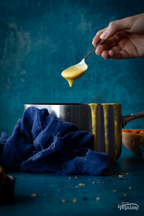 Someone reaching in a saucepan of butternut squash soup with a metal soup spoon set against a teal backdrop. There are drips of soup down the side of the pan, half a butternut squash, a pot of croutons and a blue linen napkin in the backgtround.