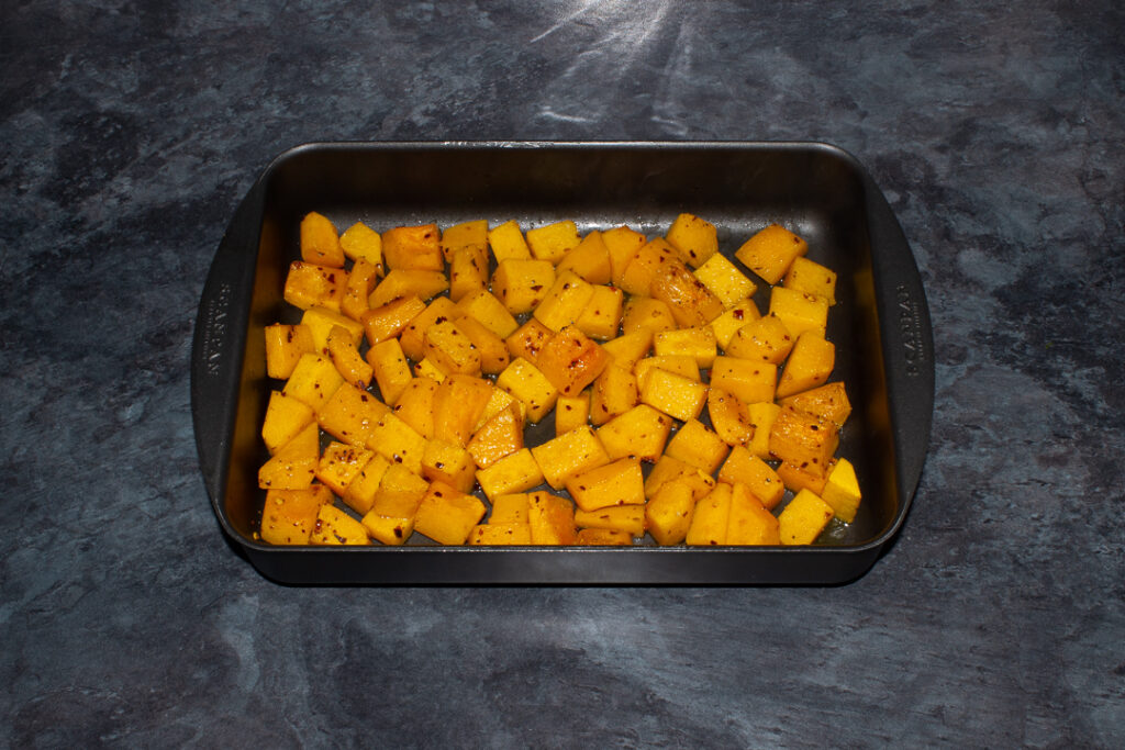 Roasted cubes of butternut squash that have been seasoned with chilli flakes in a roasting pan. Set on a kitchen worktop.