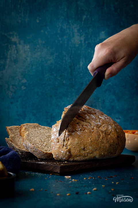 A hand reaching in to slice a loaf of sourdough bread with a bread knife on a dark wood chopping board. All set against a teal backdrop, there's also half a butternut squash, a pot of croutons, a deep blue linen napkin, chilli flakes and breadcrumbs in the background.