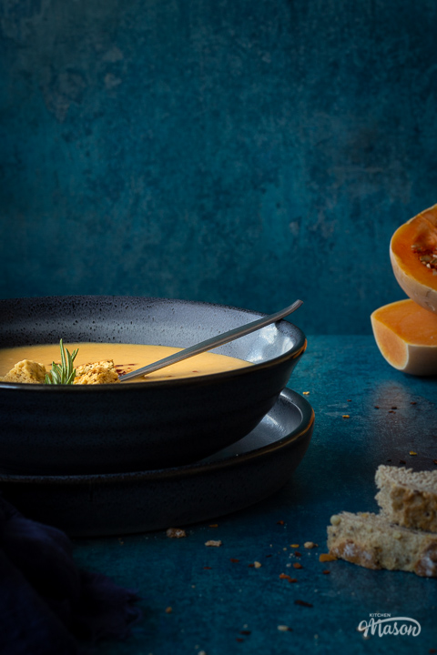 Front view of a bowl of spicy roast butternut squash soup with a spoon in it set over a plate. There is a dark blue linen napkin, two halves of a butternut squash and some bread in the background. Set on a teal backdrop.