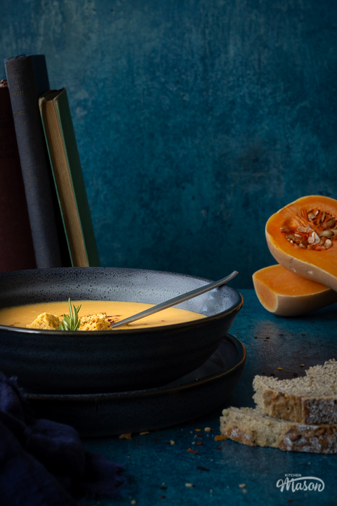 Front view of a bowl of spicy roast butternut squash soup with a spoon in it set over a plate. There is a dark blue linen napkin, some books stacked up at the back, two halves of a butternut squash and some bread in the background. Set on a teal backdrop.
