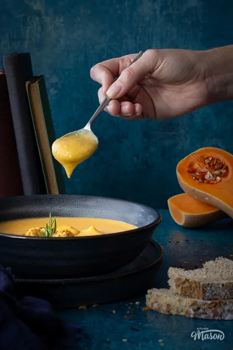 Front view of a bowl of spicy roast butternut squash soup with a hand holding up a spoon filled with soup above it set over a plate. There is a dark blue linen napkin, some books stacked up at the back, two halves of a butternut squash and some bread in the background. Set on a teal backdrop.