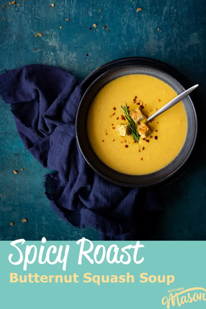 Flat lay view of spicy roast butternut squash soup with a spoon in a blue bowl set on a blue plate with a dark blue linen napkin on the side. Set on a teal backdrop, there are a few chilli flakes and breadcrumbs scattered in the background. A text overlay says "spicy roast butternut squash soup".