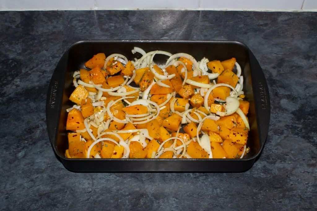 Cubes of butternut squash roasted with chilli flakes in a roasting dish. Sliced onion and garlic has been added to the pan. Set on a kitchen worktop.