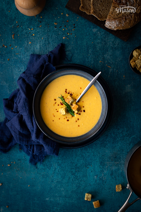 Flat lay view of spicy roast butternut squash soup with a spoon in a blue bowl set on a blue plate with a dark blue linen napkin on the side. Set on a teal backdrop, there's also a board with sliced bread, a pot with croutons, a squash and a pan with more soup in the background.