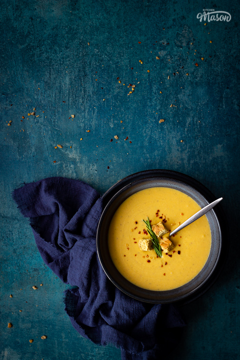 Flat lay view of spicy roast butternut squash soup with a spoon in a blue bowl set on a blue plate with a dark blue linen napkin on the side. Set on a teal backdrop, there are a few chilli flakes and breadcrumbs scattered in the background.