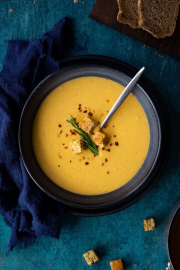 Flat lay view of spicy roast butternut squash soup with a spoon in a blue bowl set on a blue plate with a dark blue linen napkin on the side. Set on a teal backdrop, there's also a board with sliced bread, a pot with croutons and a pan with more soup in the background.