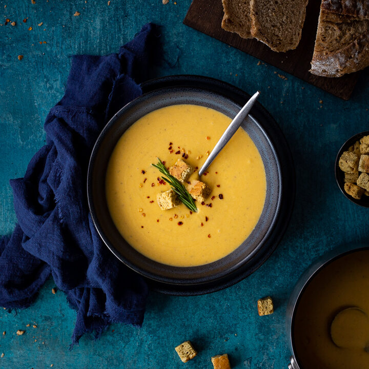 Flat lay view of spicy roast butternut squash soup with a spoon in a blue bowl set on a blue plate with a dark blue linen napkin on the side. Set on a teal backdrop, there's also a board with sliced bread, a pot with croutons and a pan with more soup in the background.