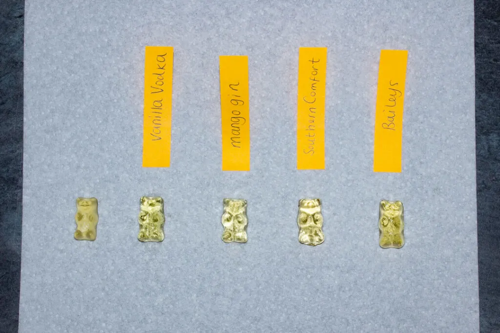 A piece of kitchen paper with an original unsoaked gummy bear on it followed by gummy bears soaked in each of the 4 tested spirits. (There are labels stating which one each gummy bear hs been soaked in.)