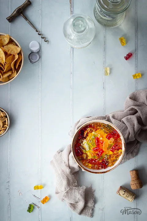 Flat lay view of vodka gummy bears in a bowl over a light brown linen napkin. There is also a bowl of crisps, a bowl of cashew nuts, 2 bottles of spirits, a cork screw, 2 bottle tops, 2 corks and gummy bears in the background. Set over a pale green wood effect backdrop.