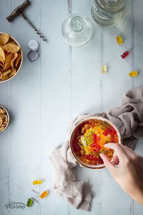 Flat lay view of a hand reaching for vodka gummy bears in a bowl over a light brown linen napkin. There is also a bowl of crisps, a bowl of cashew nuts, 2 bottles of spirits, a cork screw, 2 bottle tops, 2 corks and gummy bears in the background. Set over a pale green wood effect backdrop.