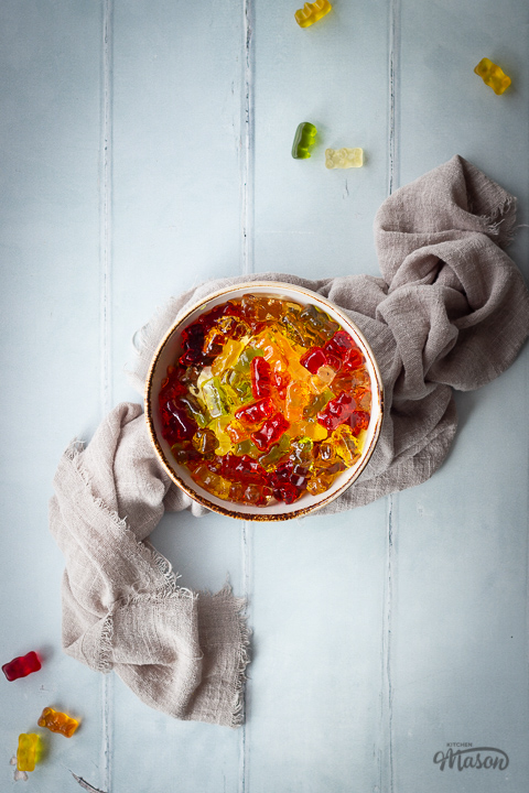 Flat lay view of vodka gummy bears in a bowl over a light brown linen napkin. Set on a pale green wood effect backdrop with gummy bears scattered in the background.