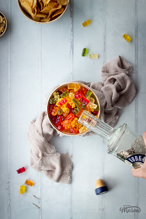 Flat lay view of vodka gummy bears in a bowl with a hand pouring vodka into them, set on a light brown linen napkin. Set on a pale green wood effect backdrop, there is also a bowl of crisps, a bowl of cashew nuts, a bottle top and gummy bears in the background.