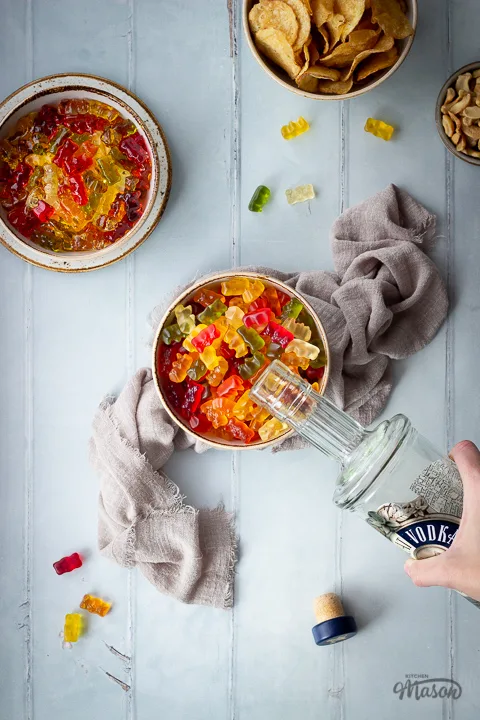 Flat lay view of vodka gummy bears in a bowl with a hand pouring vodka into them, set on a light brown linen napkin. Set on a pale green wood effect backdrop, there is also a bowl of crisps, a bowl of cashew nuts, another bowl of vodka gummy bears set on a plate, a bottle top and gummy bears in the background.