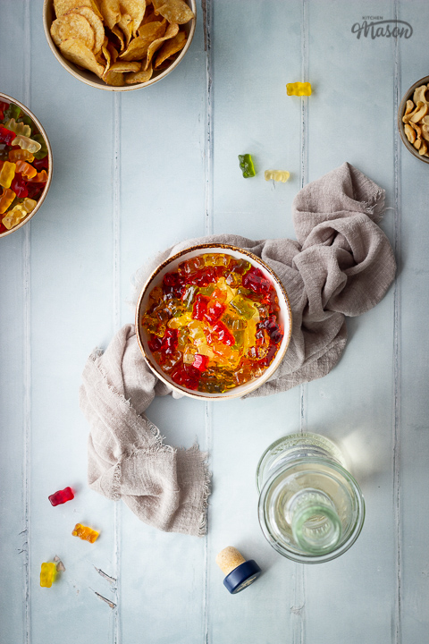 Flat lay view of vodka gummy bears in a bowl set on a light brown linen napkin. Set on a pale green wood effect backdrop, there is also a bowl of crisps, a bowl of cashew nuts, another bowl of vodka gummy bears set on a plate, an open bottle of vodka, a bottle top and gummy bears in the background.