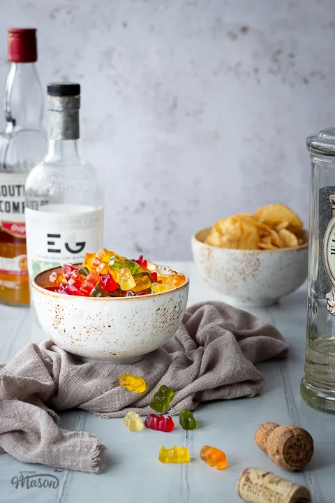 Front view of vodka gummy bears in a white bowl over a light brown linen napkin. Set over a pale green wood effect backdrop, there are also 2 bottles of spirits and a bowl of crisps in the background and a bottle of vodka, a cork and scattered gummy bears in the foreground.
