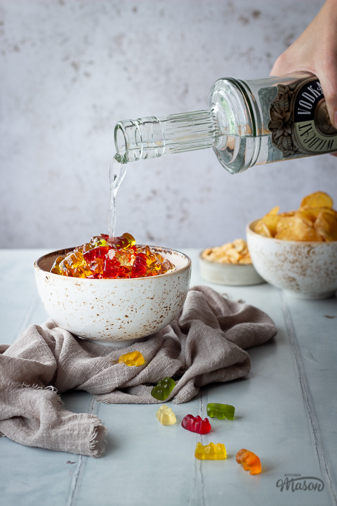 Front view of vodka gummy bears in a white bowl over a light brown linen napkin with a hand pouring vodka into them. Set over a pale green wood effect backdrop, there is also a bowl of crisps and a bowl of cashews in the background, and scattered gummy bears in the foreground.