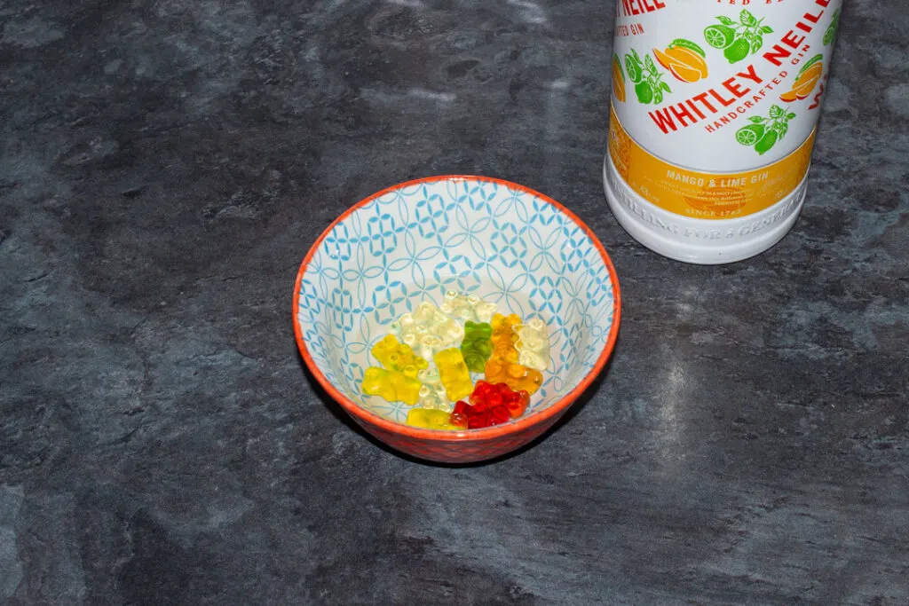 Gummy bears covered with mango and lime gin in a bowl on a kitchen worktop. There's a bottle of mango and lime gin in the background.