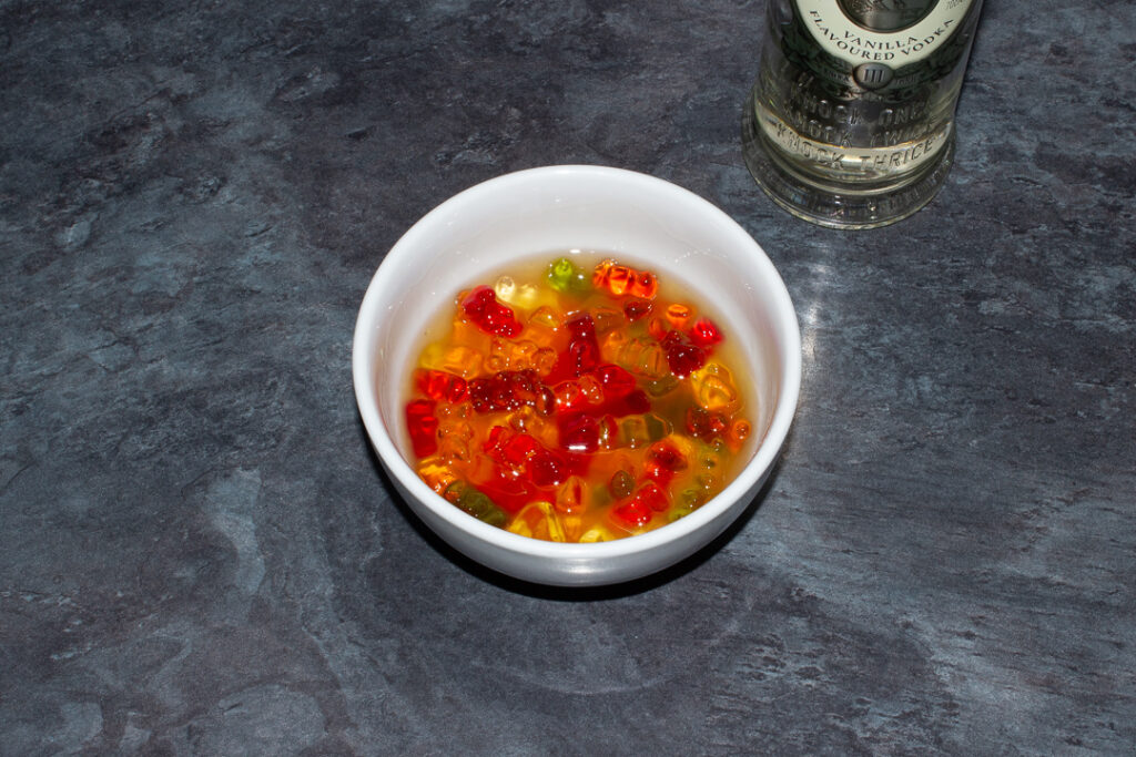 Gummy bears soaked in vodka in a white bowl on a kitchen worktop. There's a bottle of vodka in the background.
