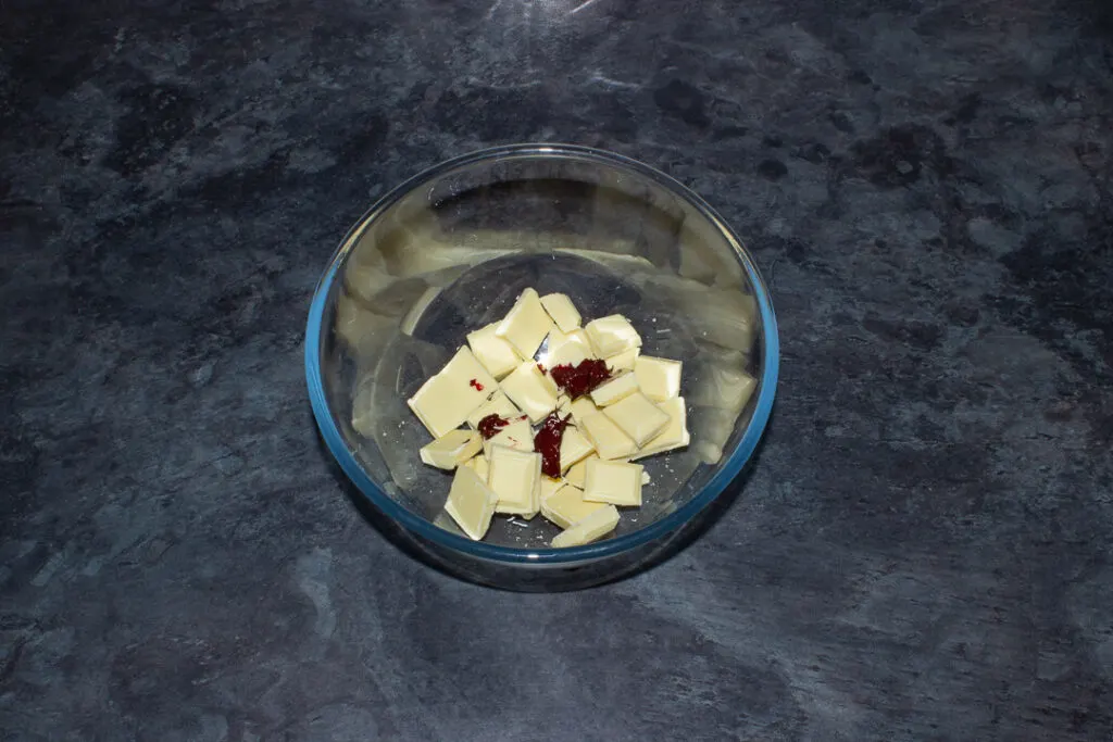 Broken white chocolate and red food colouring gel in a glass mixing bowl on a kitchen worktop.