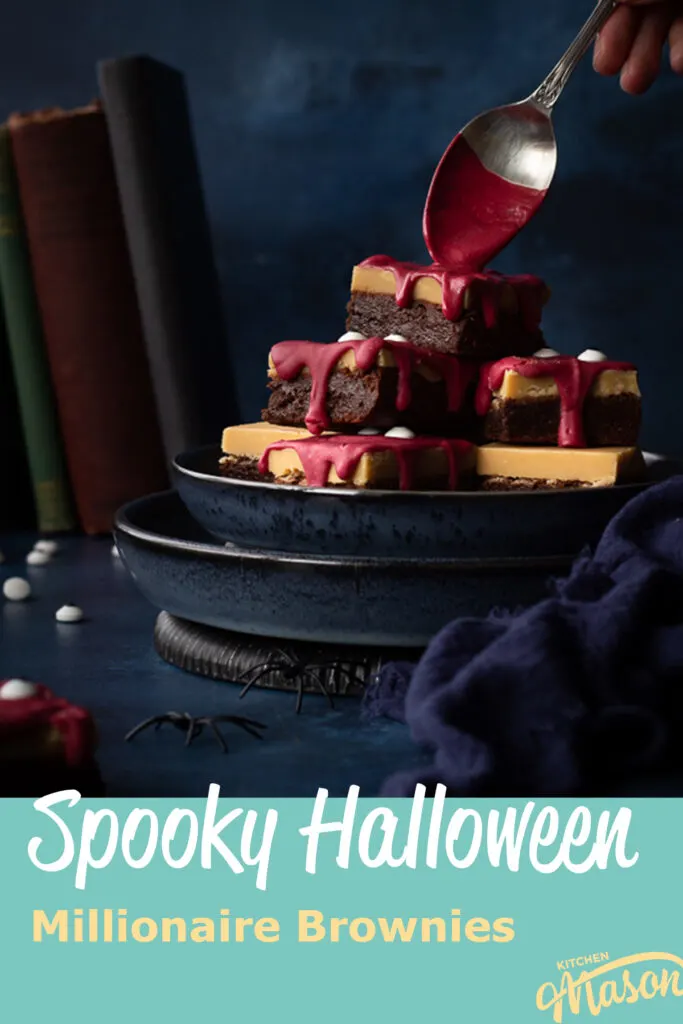 Front view of someone pouring melted red chocolate on a pyramid of Halloween Millionaire brownies, set on stacked blue plates against a deep blue hand painted backdrop. There is also a blue linen napkin, fake spiders, edible eyes and some old books in the background. A text overlay says "Spooky Halloween millionaire brownies".