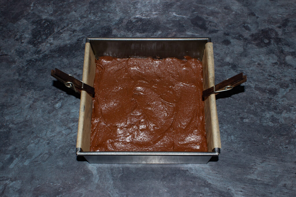Brownie batter smoothed out in a lined square baking tin on a kitchen worktop.